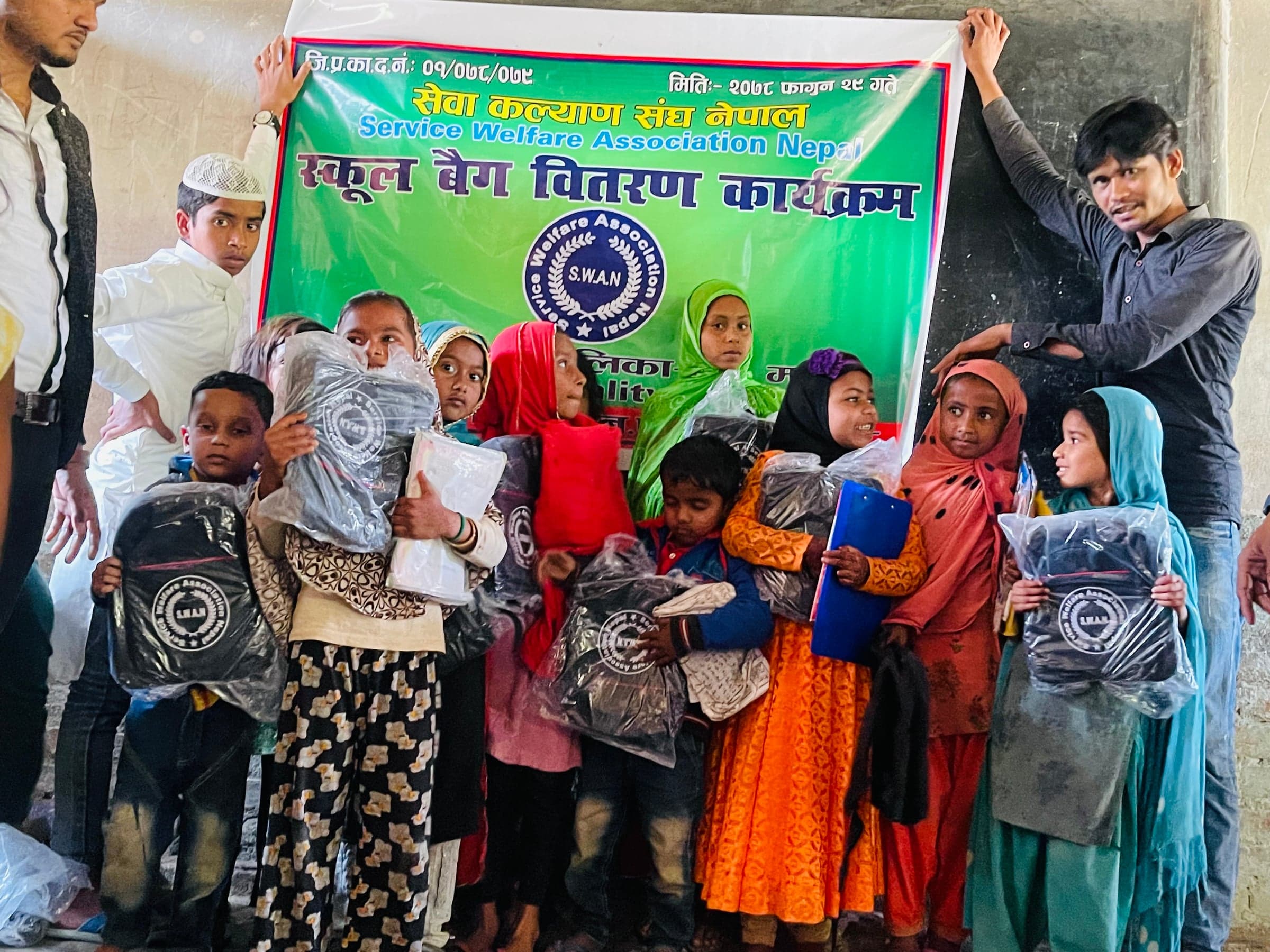 Children hold school bags and learning materials in front of a Service Welfare Association Nepal banner.